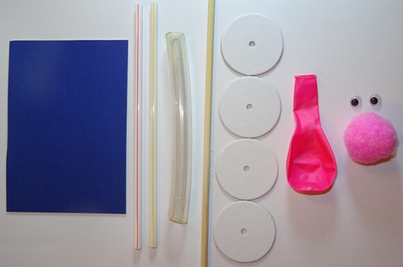 Images shows the parts needed to make a balloon buggy, featuring a piece of coloured card, 2 straws, a piece of hose, a dowel, 4 card wheels, a balloon, 2 googly eyes and a large pompom. 