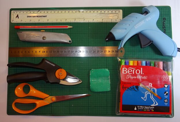 Images shows tools and consumables needed for a balloon buggy, featuring rulers, scissors, a stanley knife, a pair of secateurs, a pencil, cutting mat, sandpaper and felt tip pens. 
