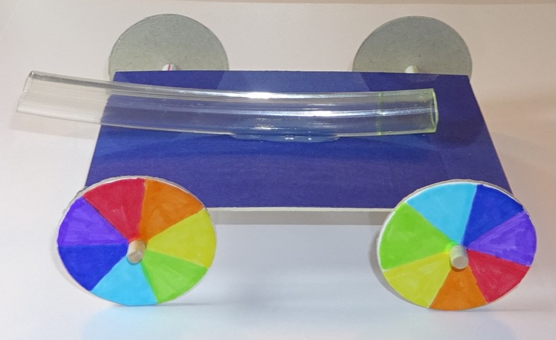 Image of a partial completed balloon buggy, showing a board with four, coloured wheels attached and a hose attached on top. 
