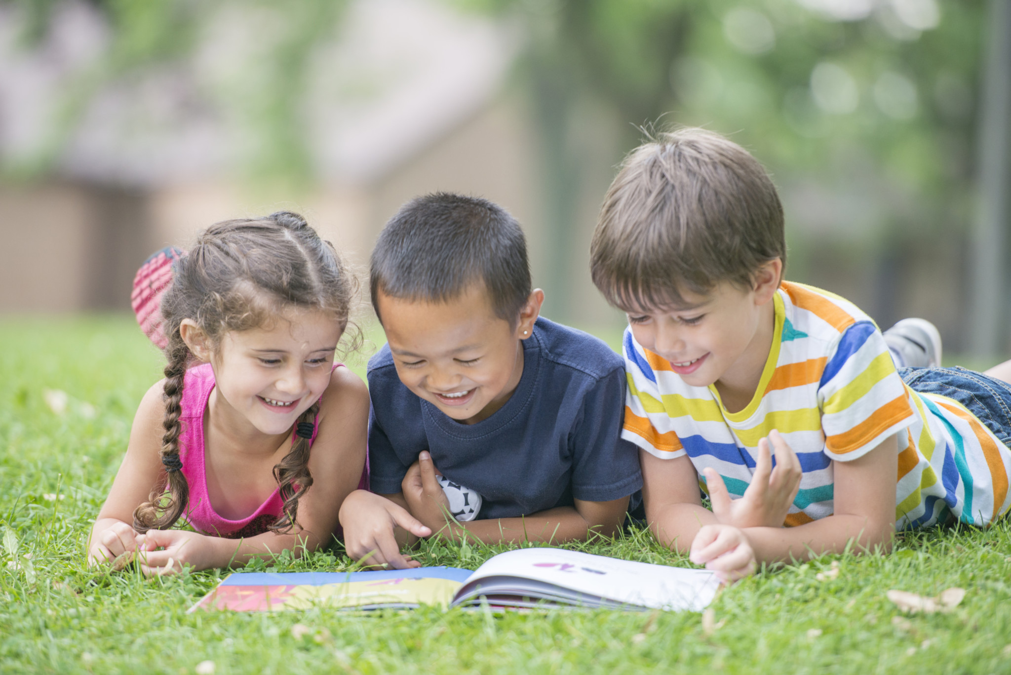 A multi-ethnic group of elementary age children are lying in the grass and are reading a book together in the grass.